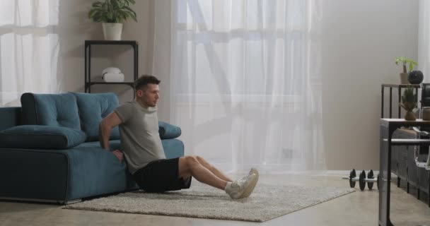 Дорослий спортивний чоловік тренується вдома, використовує меблі як тренажерне обладнання, тренується у вітальні, спирається на диван, тримає посадку — стокове відео