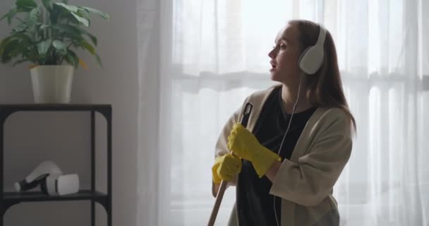 Поющая домработница танцует со шваброй в гостиной, уборка и уборка, средний портрет в помещении — стоковое видео