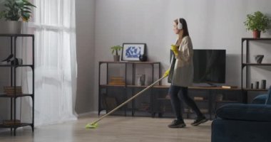 Oda temizliği ve apartman temizliği, neşeli genç kadın yerleri paspas ve dansla yıkıyor, kulaklıkla müzik dinliyor.