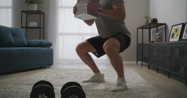Intenzivní trénink doma, člověk dřepí s vahou v obývacím pokoji, drží knihy v rukou, silové cvičení — Stock video