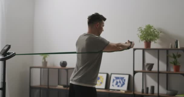 Dorosły mężczyzna z muskularnym i seksownym ciałem trenuje sam w mieszkaniu, wykonując ćwiczenia z gumką na plecy — Wideo stockowe