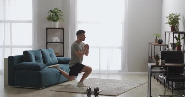 Hombre atlético guapo está entrenando en casa por la mañana, en cuclillas en una pierna, entrenamiento en la sala de estar, estilo de vida saludable — Vídeo de stock