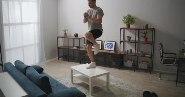 Home training met meubels in de woonkamer, man staat op tafel, fitnessruimte in appartement op zelf-isolatie, gezonde levensstijl — Stockvideo