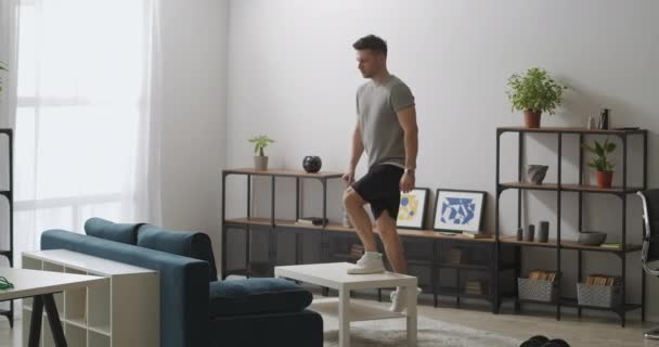 Çekici genç adam sabah oturma odasında egzersiz yapıyor, masa başı bacak kasları için egzersiz yapıyor, evdeki spor malzemeleri gibi mobilyaları kullanıyor. — Stok video