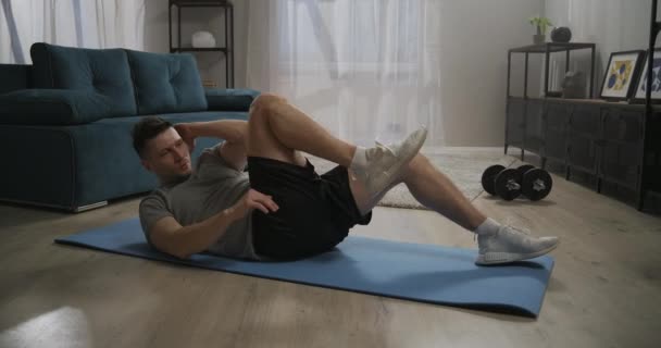 Cvičení na šikmé břišní svaly, muž trénuje doma po ránu, leží na podlaze a dělá křupky, zvedá nohy — Stock video
