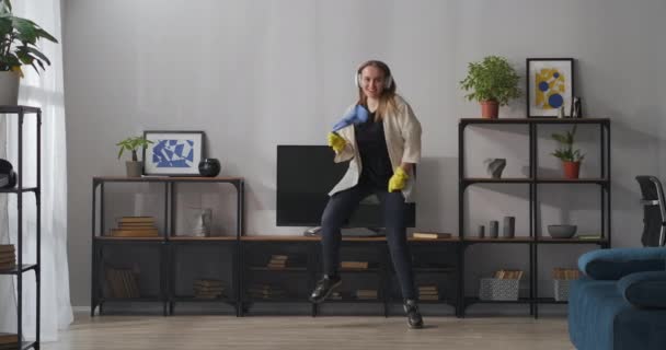 Glad galen hemmafru dansar skämtsamt i vardagsrummet, göra hushållsarbete och ha kul, lyssna på musik med hörlurar — Stockvideo