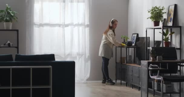 Νεαρή νοικοκυρά με ακουστικά κάνει καθαρισμό στο σαλόνι, καθαριότητα και θαλπωρή στο σπίτι, σκούπισμα σκόνη — Αρχείο Βίντεο