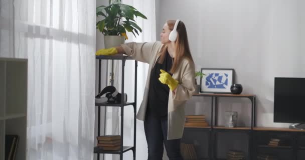 Städa hemma, kvinna lyssnar på musik med hörlurar och damning, torka hyllor av torka i modern lägenhet — Stockvideo