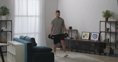 Genç sporcu hafta sonları oturma odasında tek başına antrenman yapıyor. Ağırlıklar elinde, kaslar için egzersiz yapıyor.