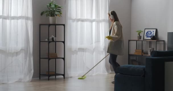 Glad och bekymmerslös kvinna lyssnar på musik under saneringen i vardagsrummet, tvätta golvet med mopp, dansa och sjunga — Stockvideo