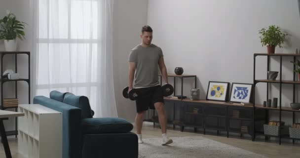 Ung sportig kille tränar ensam i vardagsrummet på helgen, huk med vikter i händerna, övningar för muskler — Stockvideo