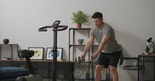Gespierde man is trainen alleen thuis, stretching elastiekje door armen, training voor een goede lichamelijke conditie en lichaamsvorm — Stockvideo