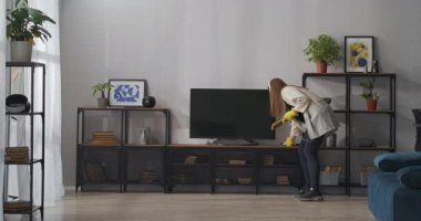 Tasasız kadın temizlik yapıyor, müzik dinliyor ve modern apartmandaki raflardan ve televizyondan toz alıyor.