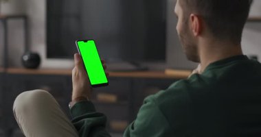 Genç adam video aracılığıyla iletişim kuruyor. Akıllı telefonuyla. Elinde krom anahtarla evde oturuyor.