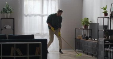 Neşeli adam evde paspas ile dans ediyor, kulaklıkla müzik dinliyor ve şarkı söylüyor, evi temizliyor.