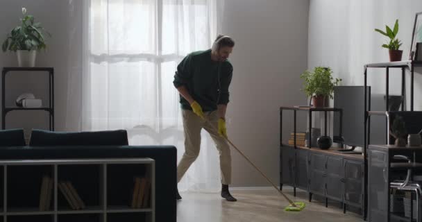 Glad man dansar med mopp i lägenheten, lyssnar på musik med hörlurar och sång, städa hemma — Stockvideo