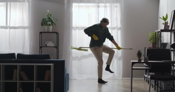 Rolig ung man dansar med mopp som gitarr och lyssnar på musik med hörlurar, städning i lägenhet på helgerna — Stockvideo