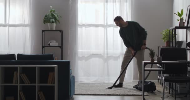 Nettoyage de la maison le week-end, l'homme est hoovering tapis dans le salon, en utilisant l'aspirateur pour l'appartement rangé à la journée ensoleillée — Video