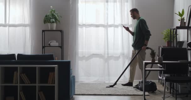 Jongeman is het reinigen met stofzuiger in appartement en surfen op internet via smartphone met wifi thuis — Stockvideo