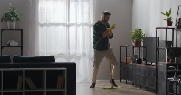 男はアパートの掃除中にモップと歌で踊ってるヘッドフォンで音楽を聴いてる — ストック動画