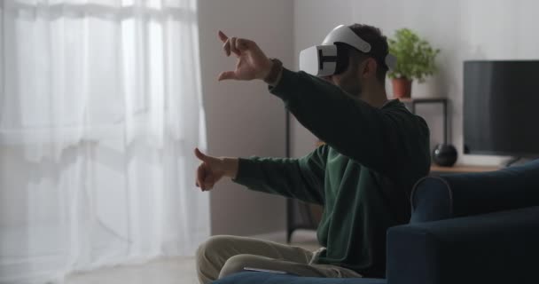 Hombre está sentado en casa y usando gafas de realidad virtual, gesticulando con las manos, deslizando y acercando imágenes — Vídeo de stock