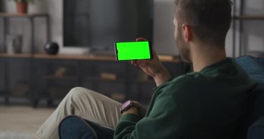 Yetişkin adam evde dinleniyor ve video konferansı ile iletişim kuruyor. Akıllı telefon ekranında, cihazda yeşil ekran.