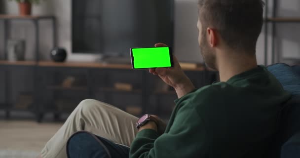 大人の男性は自宅で休んでいてスマートフォンのディスプレイ上のビデオ会議やガジェット上の緑の画面で通信しています — ストック動画