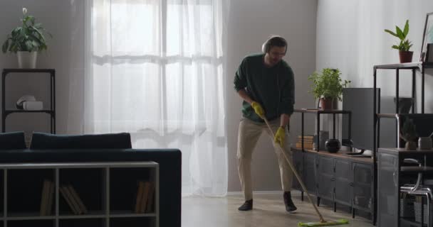 Hombre alegre está lavando piso en la sala de estar, escuchando música por auriculares y bailando divertido con fregona en casa — Vídeos de Stock