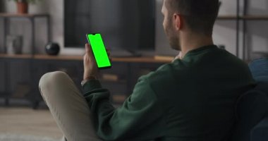 Adam internette cep telefonuyla görüntülü sohbet yapıyor, oturma odasındaki koltukta oturuyor, yeşil ekran cep telefonu, kroma anahtar teknolojisi.