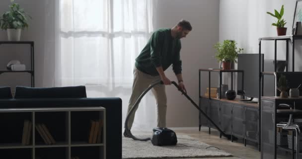 Getrouwde man maakt thuis schoon, gebruikt stofzuiger voor het reinigen van tapijt en vloer in de woonkamer — Stockvideo