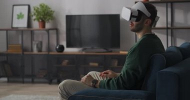 Yetişkin adam kafaya monte edilmiş ekran ve sanal gerçeklik teknolojisi kullanarak 3D video izliyor, evdeki koltukta oturuyor, modern teknoloji