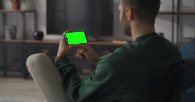 Adam evde dinleniyor. Krom anahtarı için yeşil ekranlı akıllı telefon kullanıyor. Cep telefonunu yatay olarak tutuyor.