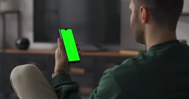 Joven hombre está sosteniendo móvil verticalmente y mirando en la pantalla, deslizando la pantalla, ver imágenes, pantalla verde para la clave de croma — Vídeo de stock