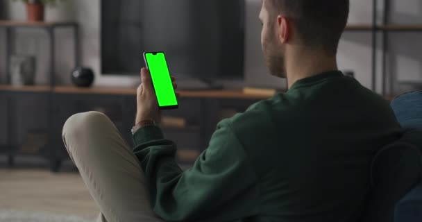 Homme communique par chat vidéo sur smartphone, assis à la maison dans le salon, se détendre sur le canapé, écran vert du téléphone portable — Video