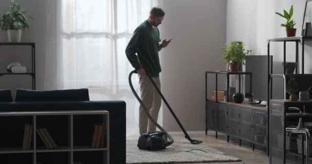 成人男性は自宅で掃除をしていますスマートフォンやインターネット中毒でソーシャルネットワークを閲覧しています掃除機で掃除機をかけています — ストック動画