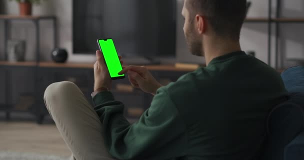 Teléfono inteligente de pantalla verde en manos del usuario masculino adulto, el hombre está sentado en el apartamento y deslizando la pantalla de los aparatos modernos — Vídeos de Stock