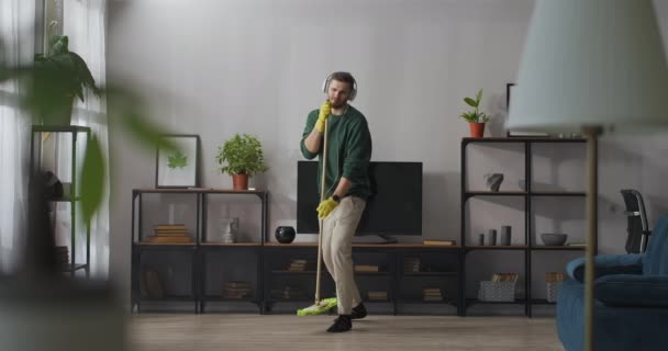 Adult handsome man is singing into mop during cleaning in apartment, listening to music in headphones, fun and amusement during home clean-up — Stock Video