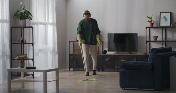 Hombre está haciendo tareas domésticas, lavando el piso en la sala de estar y escuchando música por los auriculares, ayuda alrededor de la casa — Vídeos de Stock