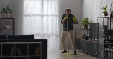 Komik adam evde temizlik yapıyor, oturma odasında yerleri yıkıyor ve mikrofon olarak paspasla şarkı söylüyor, kulaklıkla müzik dinliyor.