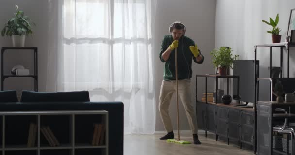 Funny guy is doing clean-up at home, washing floor in living room and singing at mop as microphone, listening to music by headphones — Stock Video