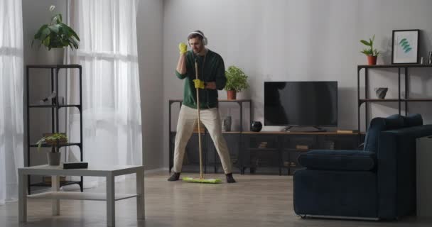 Hombre alegre está haciendo tareas domésticas, lavando el piso en la sala de estar y cantando en fregona, escuchando música por auriculares — Vídeo de stock