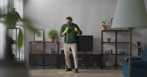 Comical man is singing into mop during clean-up in flat, listening to music in headphones, imaging himself as superstar — Stock Video