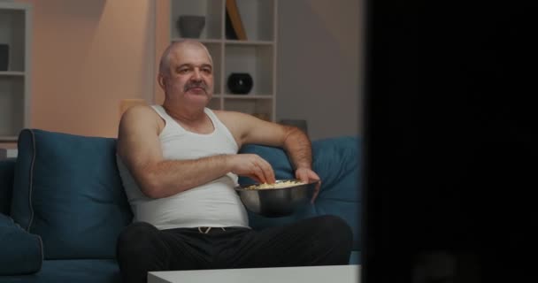 Wieczorem mężczyzna ogląda telewizję. Siedzi na kanapie. Jedzenie popcornu z dużej blachy żelaznej. kamera porusza się od lewej do prawej — Wideo stockowe