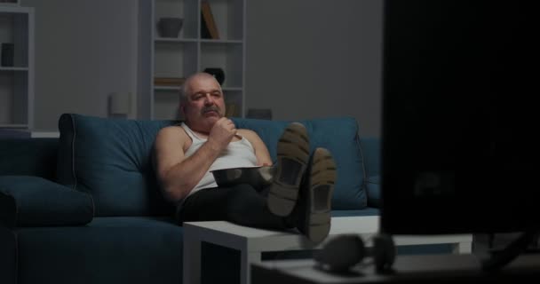 En man med mustasch tittar på kvällsnyheterna på TV och äter popcorn. Vänder huvudet åt vänster och fortsätter titta på TV. — Stockvideo