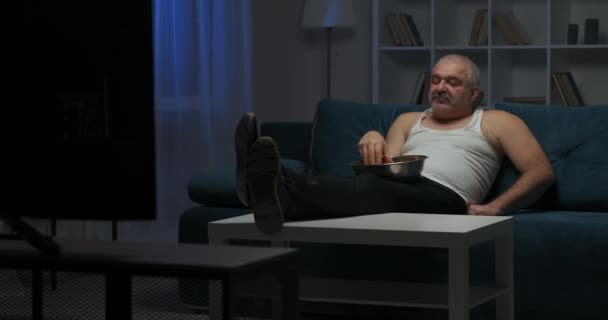 Человек с седыми короткими седыми волосами смотрит боевик по телевизору. Он ест попкорн с большой тарелки правой рукой, в то время как его левая рука лежит на диване. — стоковое видео