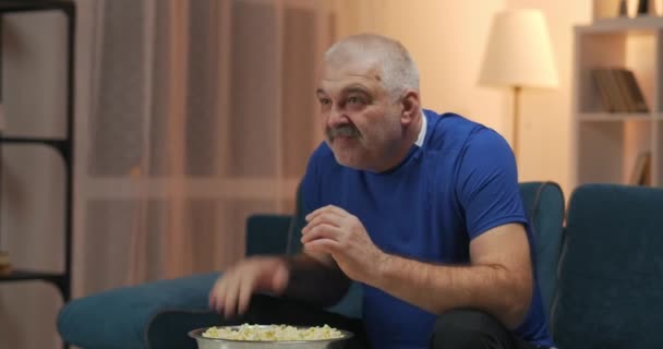 Viejo viendo un partido de deportes en la televisión. Sentado en el sofá con un plato de palomitas de maíz frente a él. Tensamente golpea la pierna con la palma de la mano, se regocija con las manos en alto. — Vídeo de stock