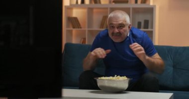 Mavi tişörtlü, bıyıklı gri saçlı bir adam televizyonda bir spor maçı izliyor. Önünde bir tabak patlamış mısırla kanepede oturmak. Olaylara duygusal tepkiler, hayal kırıklığı.