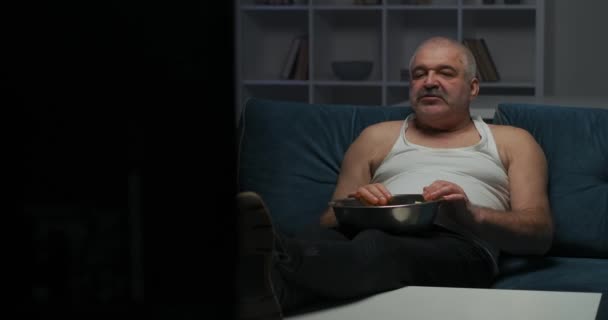 Een man met grijs kort grijs haar kijkt naar een actiefilm op tv en eet popcorn en legt zijn hand op zijn buik. Heldere flits op het scherm. — Stockvideo