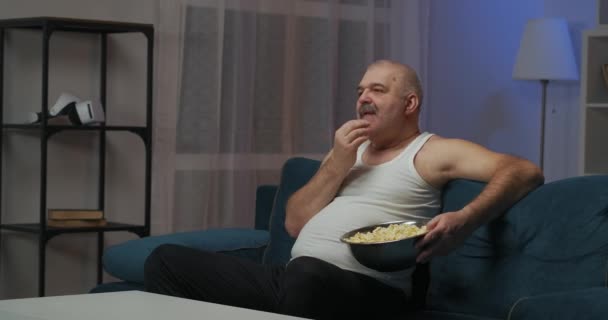 Un uomo guarda la TV la sera. E 'seduto sul divano. Mangiare popcorn da una grande piastra di ferro. la fotocamera si sposta da destra a sinistra — Video Stock