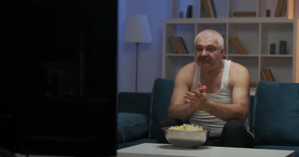 En man i vit tröja tittar på TV på kvällen medan han sitter på soffan. Äter popcorn, slår handen i knät och gnider händerna mot varandra.. — Stockvideo